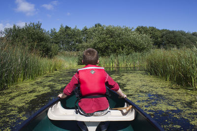 Rear view of boy sitting in boat on river against sky