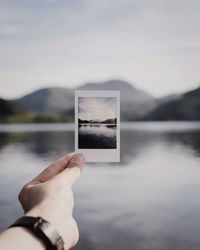 Cropped hand holding photograph against lake and mountains