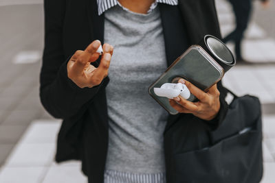 Midsection of businesswoman holding smart phone and wireless in-ear headphones while standing at street