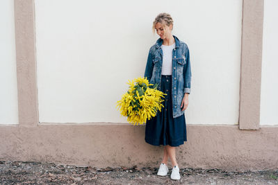 Stylish attractive white woman standing by the wall holding fresh spring fresh bouquet of mimosa.