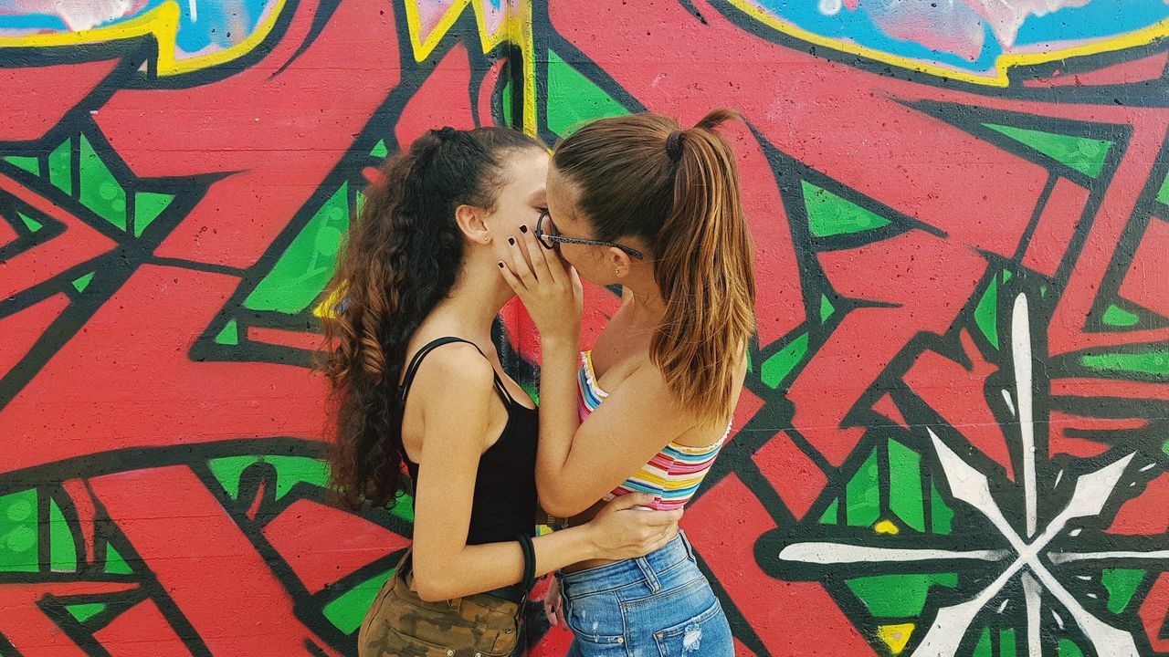 graffiti, leisure activity, women, wall - building feature, standing, lifestyles, young women, casual clothing, two people, hair, real people, young adult, hairstyle, people, wall, adult, art and craft, three quarter length, love, beautiful woman, positive emotion