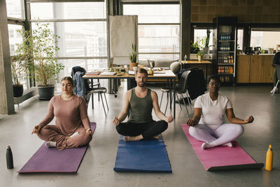 Full length of multiracial colleagues meditating together on mats at office