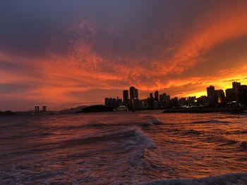 Sea by buildings against cloudy sky during sunset