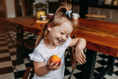 Funny little girl child in pajamas holding and eating an apple and laughing in the kitchen at home