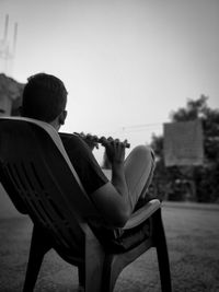 Rear view of man sitting on chair playing flute against clear sky