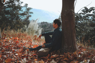 Young man looking away while sitting by tree trunk