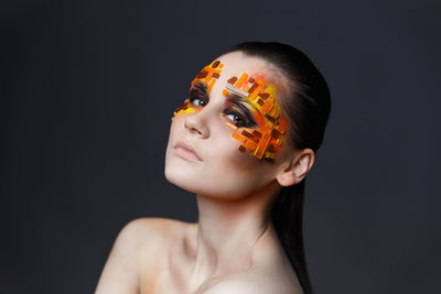 Close-up of shirtless young woman with eye make-up and rhinestones against gray background