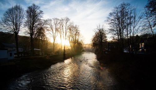 Canal amidst bare trees against sky during sunset