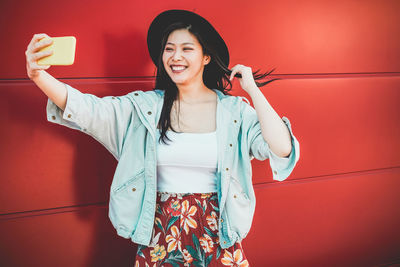 Smiling young woman taking selfie with smart phone while standing against red wall