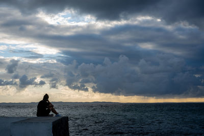 Man sitting by sea against sky during sunset