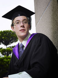 Portrait of university student wearing graduation down against column