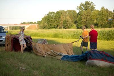 Boys assisting pilot in preparing hot air balloon on grassy field against sky during sunset