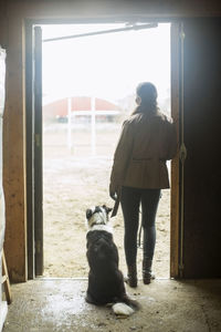 Full length rear view of young woman with dog leaning in doorway of horse stable