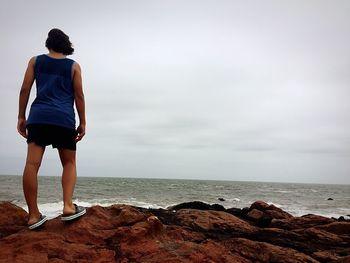 Rear view of woman standing at rocky shore against cloudy sky