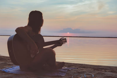 Rear view of woman sitting at beach against sky during sunset