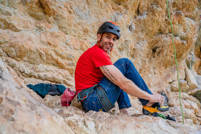 Side view of happy male alpinist looking at camera while sitting and wearing shoes on rocks