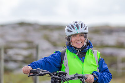 Smiling mature female cyclist with a blue jacket, helmet and anti-reflective vest next to a bicycle