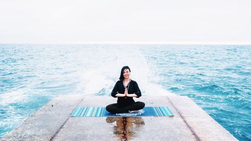 Woman in prayer position exercising on pier at sea against clear sky