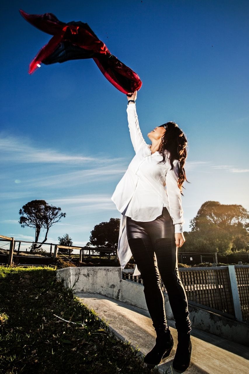 full length, lifestyles, casual clothing, leisure activity, sky, mid-air, jumping, arms outstretched, sunlight, blue, young adult, clear sky, freedom, day, low angle view, standing, skill, rear view