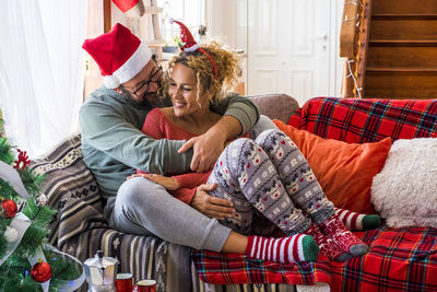 Smiling couple embracing while sitting on sofa at home by christmas tree