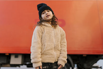 Cute girl wearing knit hat and sweater staring while standing against red truck