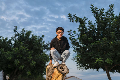 Young transgender man with glasses posing on a log outdoors.