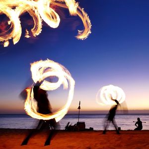 Silhouette people dancing with fire on beach during sunset