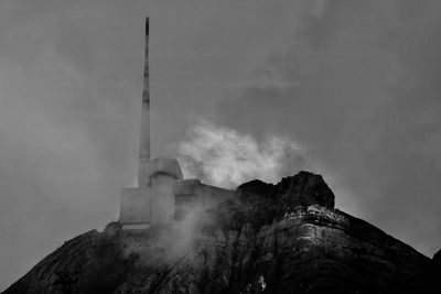 Low angle view of factory on rocky mountain against cloudy sky