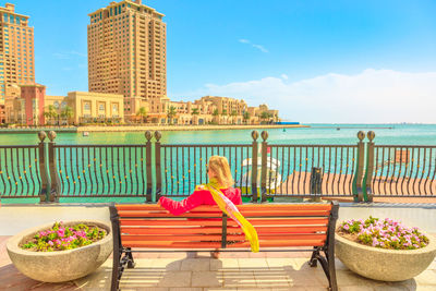 Woman sitting on bench against sea in city