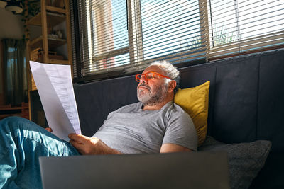 Handsome mature man wearing reading glasses sitting on sofa looks at documents while works at home.