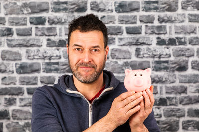 Portrait of man holding piggy bank against brick wall