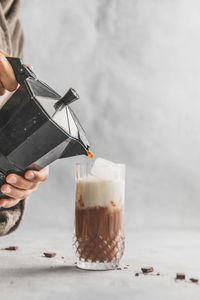 Female pouring coffee with moka pot coffee maker into dinking glass with iced mocha on table