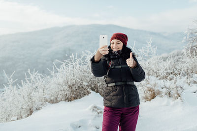 Smiling woman in warm taking selfie while standing on snowcapped mountain