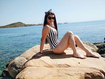 Portrait of young woman in bodysuit sitting on rock by sea