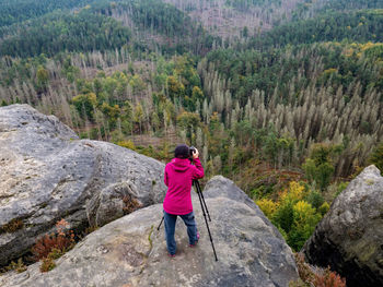 Woman photographer set tripod with camera on rock above deep valley with death dry spruce in forest