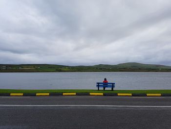 Bench on road by lake against sky