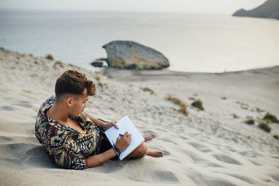 Young man sitting on sand dune while drawing in book at almeria, tabernas desert, spain