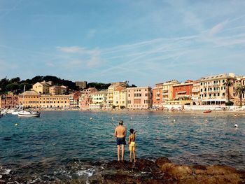 Father and son fishing in sea against buildings