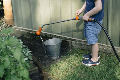 Low section of boy pouring water with garden hose in bucket at yard