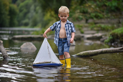 Happy little boy in yellow rain boots playing with ship boat in lake on spring or autumn day