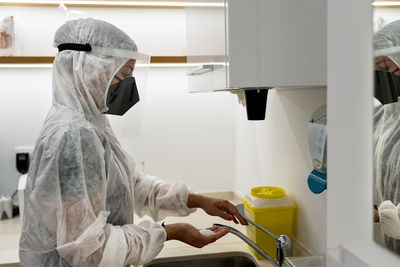 Side view of female doctor in protective suit and face shield with mask washing hands while preparing for medical procedure in modern clinic