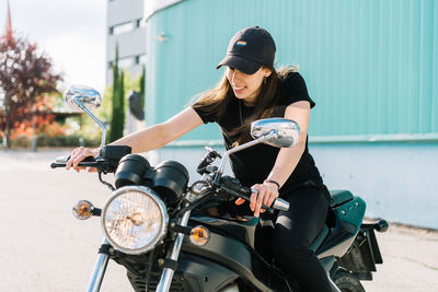 Positive female motorcyclist sitting on motorbike parked in city street on sunny day and looking away<