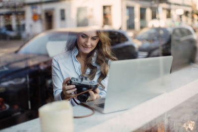 Young woman holding camera while sitting with laptop at cafe seen through window glass