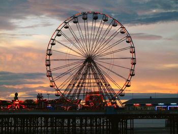 Low angle view of illuminated ferris wheel against cloudy sky at dusk