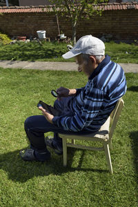 Side view full length of man using phone while sitting on chair at yard