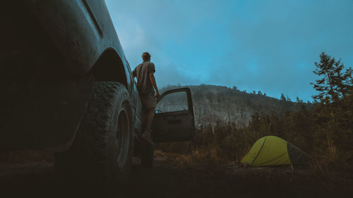 Man standing on car by tent against sky at dusk