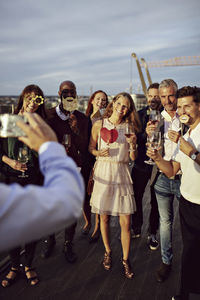 Businessman photographing happy coworkers holding wineglasses and props at office terrace in party