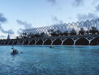 People rowing boats on sea by modern bridge against sky at dusk