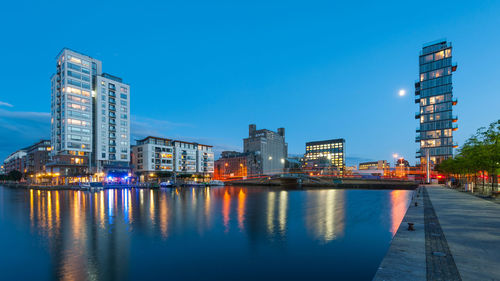 Illuminated buildings by river against sky at dusk