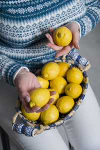 A woman in a blue sweater holds a basket with ripe yellow lemons in her hands